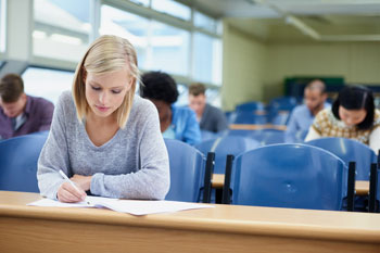 Heilpraktikerschülerin, die in einem Klassenraum sitzt und eine Prüfung schreibt. Im Hintergrund weitere Schüler/innen.